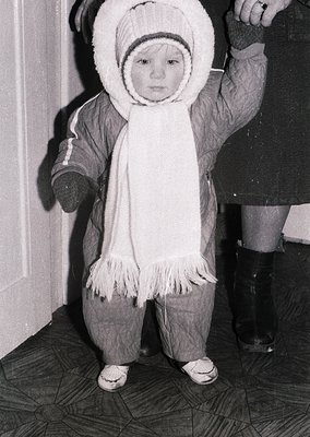 Young child in winter attire—hooded parka with fur trim, scarf, and knee-high boots—standing indoors beside an adult’s leg. M...