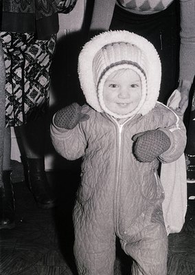 Young child in vintage winter attire, mid-20th century—puffy hooded snowsuit with mittens, posed indoors near patterned fabri...