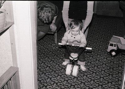 Vintage black-and-white photo of a toddler in a homemade push toy resembling a toy truck, guided by adults in a patterned car...