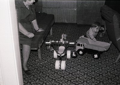 Vintage black-and-white interior shot of a woman kneeling beside a child playing with mid-century toy vehicles on patterned c...