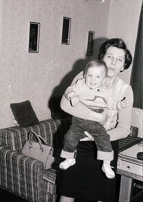 A woman cradles a toddler in a mid-century living room, likely 1950s–1960s. The child wears a striped sweater and cuffed jean...