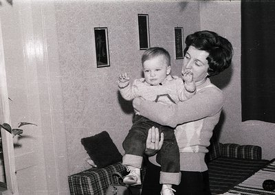 A woman in 1960s-era sweater and jeans holds a toddler on her lap in an indoor setting. Both wear layered clothing, suggestin...