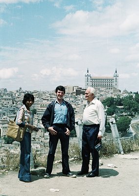 Three individuals pose outdoors in 1970s Madrid, Spain, against the backdrop of the Alcázar of Toledo. The woman wears wide-l...