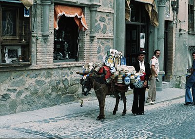Vintage street scene featuring a donkey laden with colorful crates and sacks, likely transporting goods in a historic Europea...