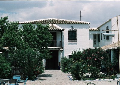 Classic Mediterranean courtyard house with stucco walls, terracotta-tiled roof, and wrought-iron balcony. Lush greenery and f...