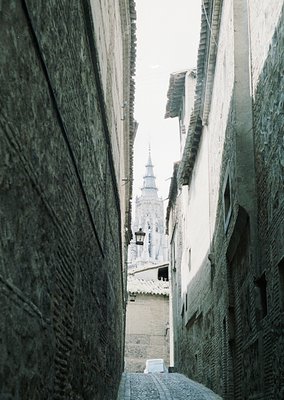 Perspective shot of a narrow, winding alley framed by aged stone walls. A historic tower with a spire rises in the distance, ...