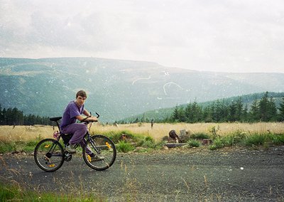 Mountain biker pauses on gravel path amid rolling hills, wearing purple long-sleeve shirt and yellow knee pads. Overcast skie...