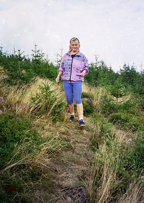 Woman in 1970s-style outdoor gear—purple jacket, high-waisted trousers, and knee-high socks—walks along a grassy, unpaved tra...