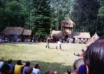 Traditional thatched huts and wooden structures in a forested setting, likely a cultural festival or reenactment. Central fig...