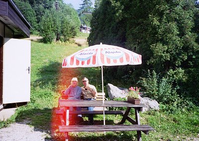 Two individuals relax under a vintage **Alpina** red-and-white striped umbrella at a rustic picnic table in a lush alpine set...