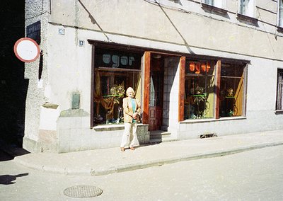Retro storefront with Cyrillic sign "АГОРА" (Agora) in a European urban setting, likely 1970s-80s. Woman in light-colored blo...
