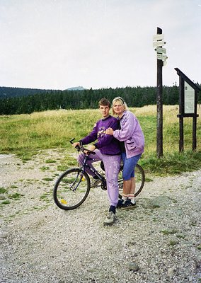 Two individuals pose on a gravel path in a forested, mountainous area, likely . The man stands on a purple mountain bike, whi...