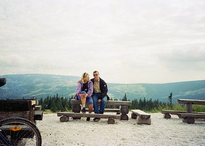 Couple posing on wooden benches atop a forested mountain ridge, 1990s-era clothing. Scenic backdrop of dense coniferous fores...