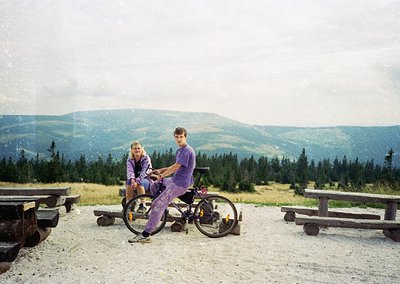 Two individuals pose on a gravelled overlook with mountain views, wearing matching purple cycling attire. The man holds a vin...