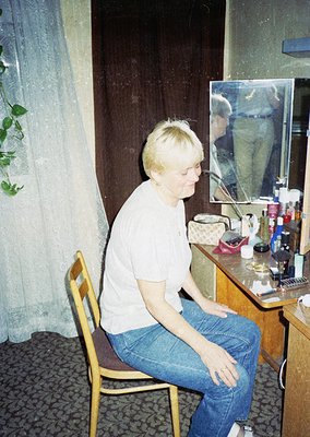 A person sits on a wooden chair in a vintage bathroom, adjusting their hair in front of a mirror with a small framed photo. W...
