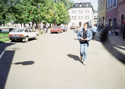 Young man in 1980s/90s streetwear—denim jacket, jeans, and sneakers—walks along a European urban street lined with parked car...