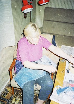 Vintage indoor portrait of a person sewing by hand on a wooden table, surrounded by fabric scraps and a sewing kit. The setti...