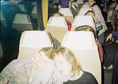 Vintage bus interior with passengers seated in rows of padded, beige upholstered seats. Blurred faces suggest motion or low-r...