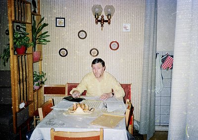 Vintage indoor dining scene featuring a man seated at a table covered with a floral-patterned tablecloth, surrounded by vinta...