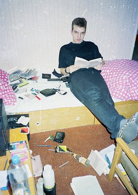 Young man reading in a cluttered, minimalist bedroom—1990s/early 2000s aesthetic. Notebooks, CDs, and a camera lie scattered ...