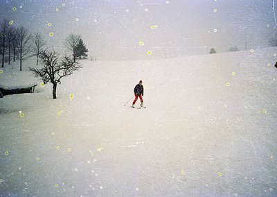 A lone skier in classic alpine gear descends a snow-covered slope during heavy snowfall, with sparse trees lining the backgro...