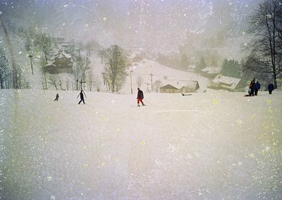 Winter scene featuring cross-country skiers navigating a snow-covered landscape. Snow-laden trees and rustic wooden buildings...