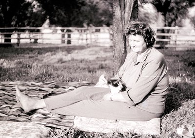 Mid-20th century woman reading outdoors on a striped blanket, holding a small white-and-brown dog. Wooden fence and rural lan...