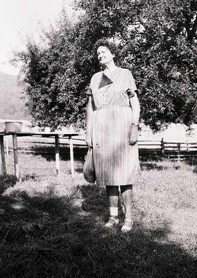 Mid-century outdoor portrait of woman in a striped, knee-length dress with a geometric collar, standing on grass beside a woo...