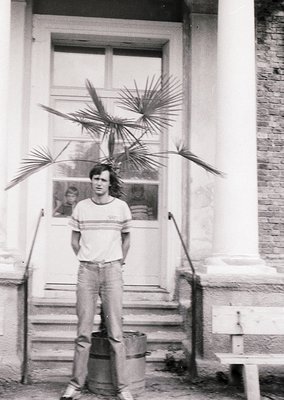 Young man in 1970s-style striped polo shirt and baggy jeans poses on brick steps beside a potted palm plant. Plain brick buil...