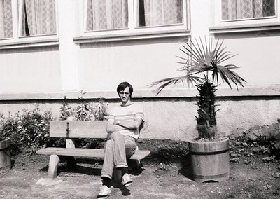 Black-and-white portrait of a person seated on a simple wooden bench in an urban courtyard. The individual wears a striped sh...