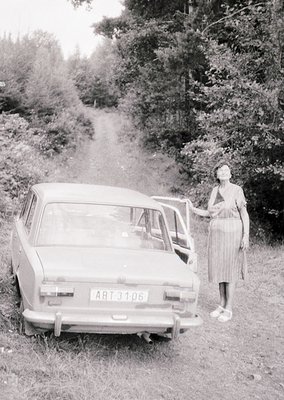 Vintage sedan (ART 3106) parked on a rural dirt road, surrounded by dense forest. Woman in 1960s-style dress poses beside it....