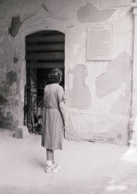 Mid-20th century girl in pleated dress stands at weathered stone doorway with wrought-iron gate. Plaque on wall suggests hist...
