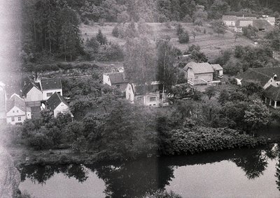 Aerial black-and-white photo of a lakeside village, likely mid-20th century. Clustered houses with pitched roofs and dense gr...