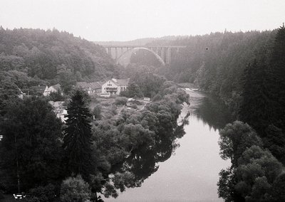 Vintage black-and-white aerial view of a grand arched viaduct spanning a river, flanked by dense forest. Mid-20th century ind...