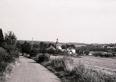 Black-and-white rural scene featuring a narrow, paved road flanked by overgrown vegetation. In the distance, a church with a ...