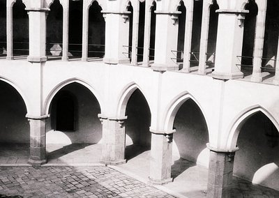 Gothic-style cloister arcade with pointed arches, stone columns, and metal railings. Likely European monastic or university a...