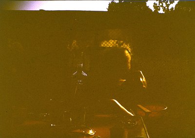Vintage low-light portrait of a person wearing a patterned headscarf, seated indoors near a drum set. Warm sepia tone suggest...