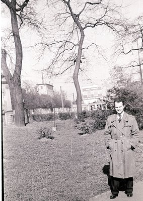 Mid-century man in a long overcoat poses outdoors in a park-like setting, likely 1950s–1960s. Bare trees and a distant indust...