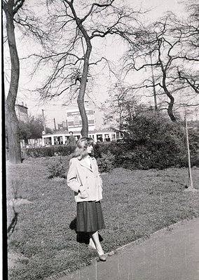Mid-century woman in a light blazer and pencil skirt strolls along a paved path in a park, flanked by bare winter trees. Urba...