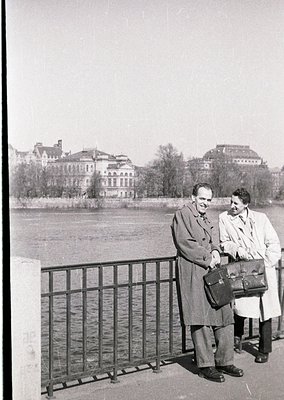 Two men in mid-20th century attire pose by a riverbank railing, one holding a briefcase. Behind them, grand neoclassical buil...
