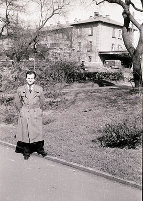 Mid-century man in a belted trench coat poses on a tree-lined urban sidewalk, 1950s–60s. Multi-story residential building wit...