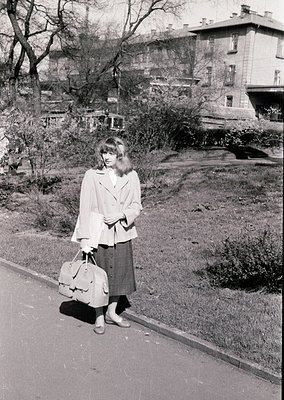 Mid-century urban portrait: Woman in 1960s streetwear—pleated skirt, cardigan, and handbag—stands on a paved sidewalk beside ...
