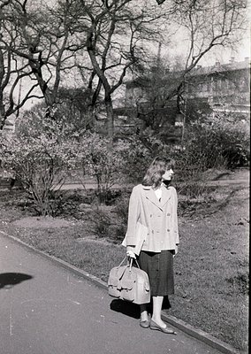Mid-century street portrait: Woman in a structured blazer, pleated skirt, and knee-high socks stands on a paved path, holding...