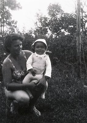A woman and infant pose outdoors in a lush, wooded area, likely mid-20th century. The woman wears a floral-patterned dress wi...