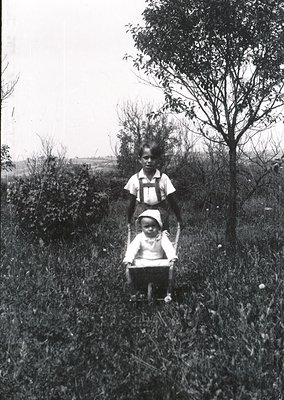 Mid-20th century black-and-white photo of two children in rural outdoor setting. Older child pushes a baby buggy with younger...