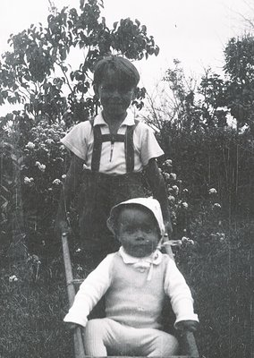 Vintage black-and-white portrait of a young boy standing, holding an infant seated on a bench in an outdoor setting with dens...