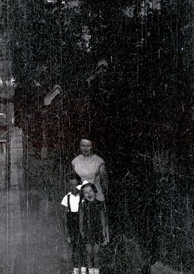 Vintage black-and-white photo of two children in rural forest setting, mid-20th century. Girl in long dress and headscarf lea...