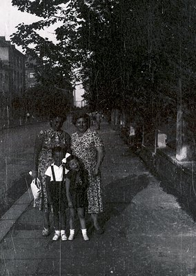 Family portrait in mid-20th century urban setting, likely 1950s–1960s. Three adults and two children pose on a tree-lined sid...