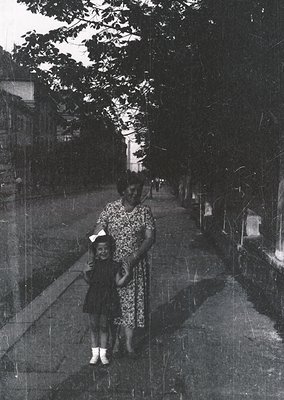 A mid-20th century black-and-white photo of a woman and child walking on a tree-lined path, likely 1950s–1960s. The woman wea...