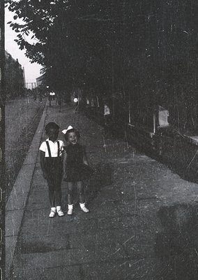 Two children pose on a wide, tree-lined concrete pathway, likely mid-20th century. Girl in a knee-length dress with white soc...